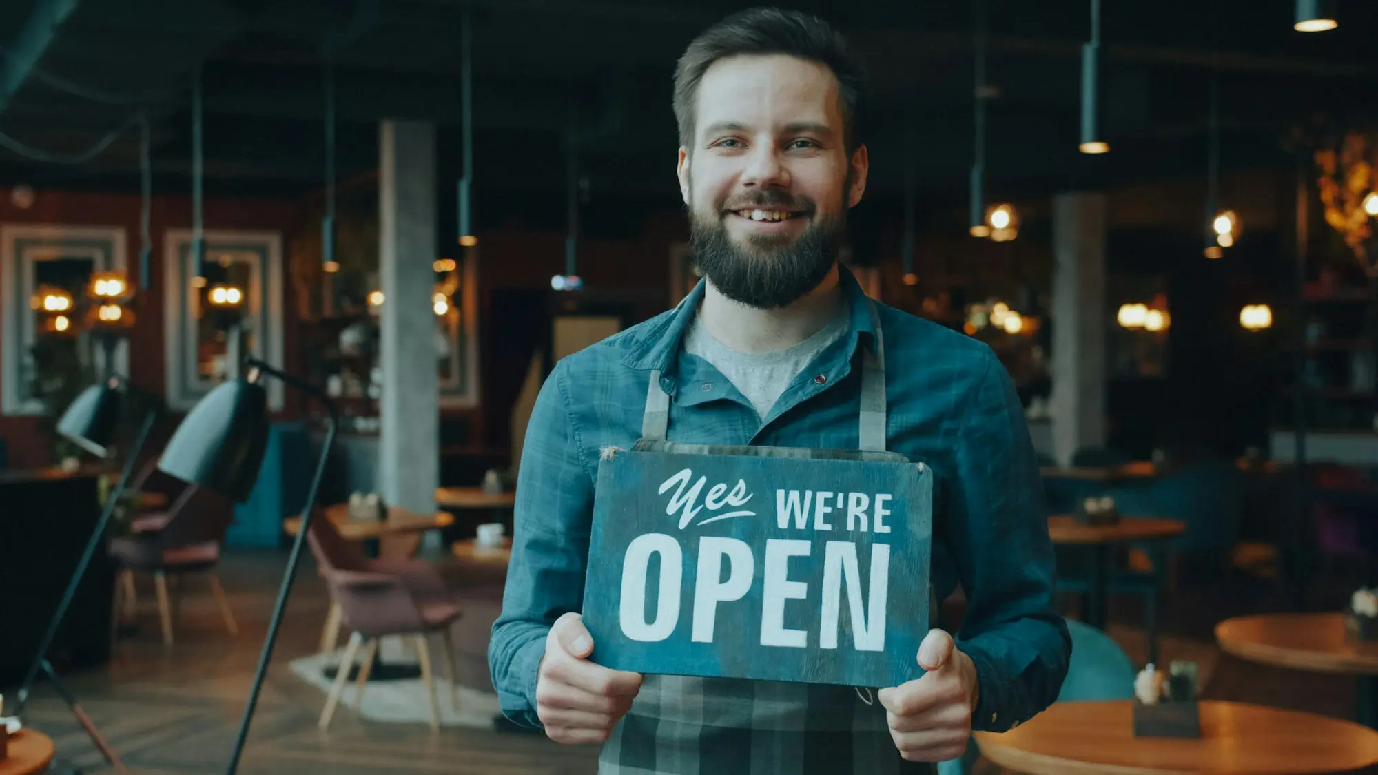 Smiling café owner holding a We're Open sign in his shop doorway
